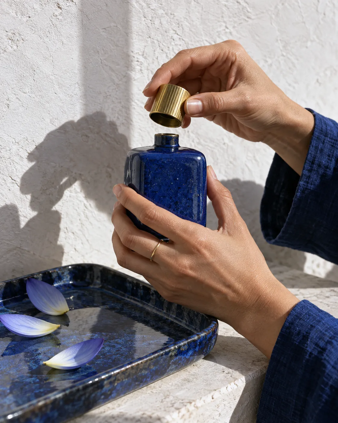 Hands lifting the gold cap off a blue faience flacon over a lapis-blue tray of lotus petals